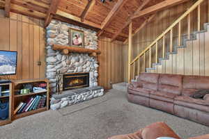 Carpeted living area with wooden walls, a stone fireplace, wood ceiling, and vaulted ceiling with beams