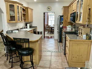 Kitchen featuring glass insert cabinets, black appliances, a kitchen breakfast bar, light tile patterned floors, and light wood finish cabinets