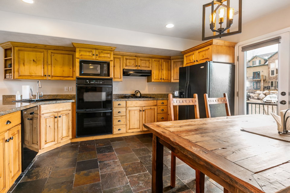 Kitchen with black appliances, stone tile flooring, dark countertops, open shelves, and wood finish cabinets