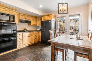 Kitchen with black appliances, light wood finish cabinetry, a chandelier, and stone tile floors