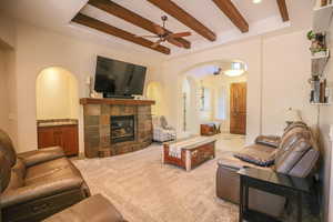 Living room featuring arched walkways, a glass covered fireplace, a ceiling fan, beam ceiling, and light carpet