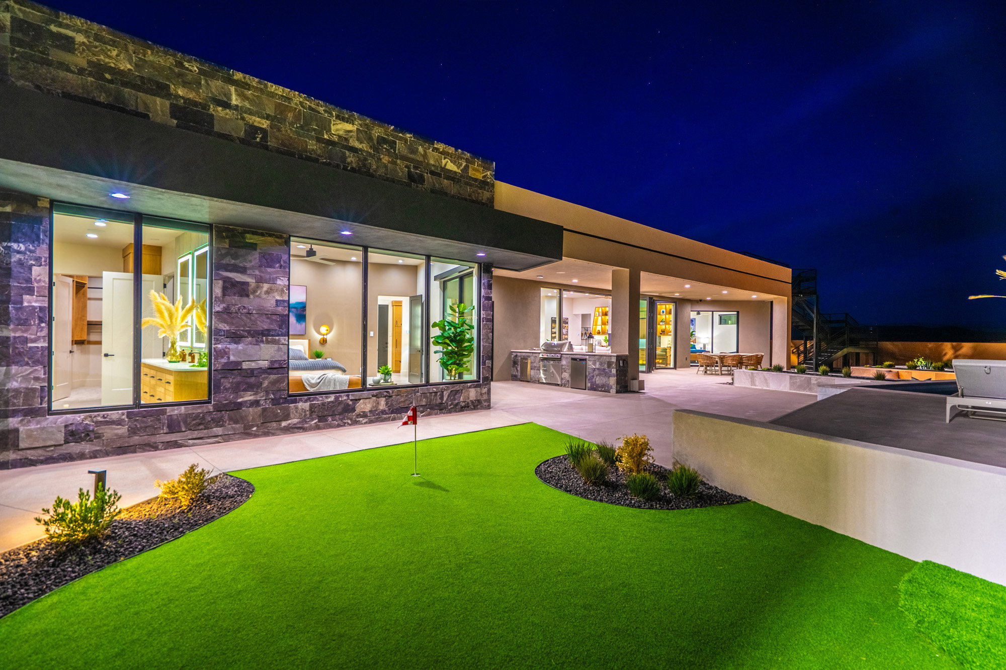 Back of house at night featuring an outdoor kitchen, an area to practice putting, a patio, and stone siding