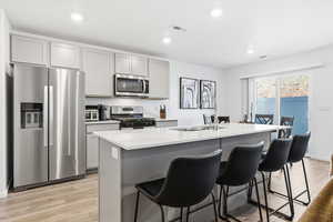Kitchen with stainless steel appliances, gray cabinetry, light wood finished floors, a kitchen island with sink, and a breakfast bar area