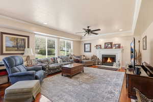 Living room featuring light wood-style flooring, ceiling fan, a warm lit fireplace, crown molding, and recessed lighting