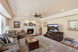 Living room with a warm lit fireplace, a ceiling fan, wood finished floors, ornamental molding, and recessed lighting