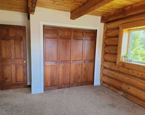 Unfurnished bedroom with rustic walls, a wood ceiling with exposed beams, carpet flooring, and a closet
