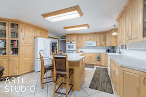 Kitchen featuring glass fronted cabinets, a breakfast bar area, backsplash, white appliances, and light tile patterned floors