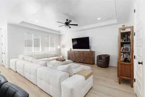 Living area featuring a raised ceiling, light wood-type flooring, ceiling fan, and recessed lighting