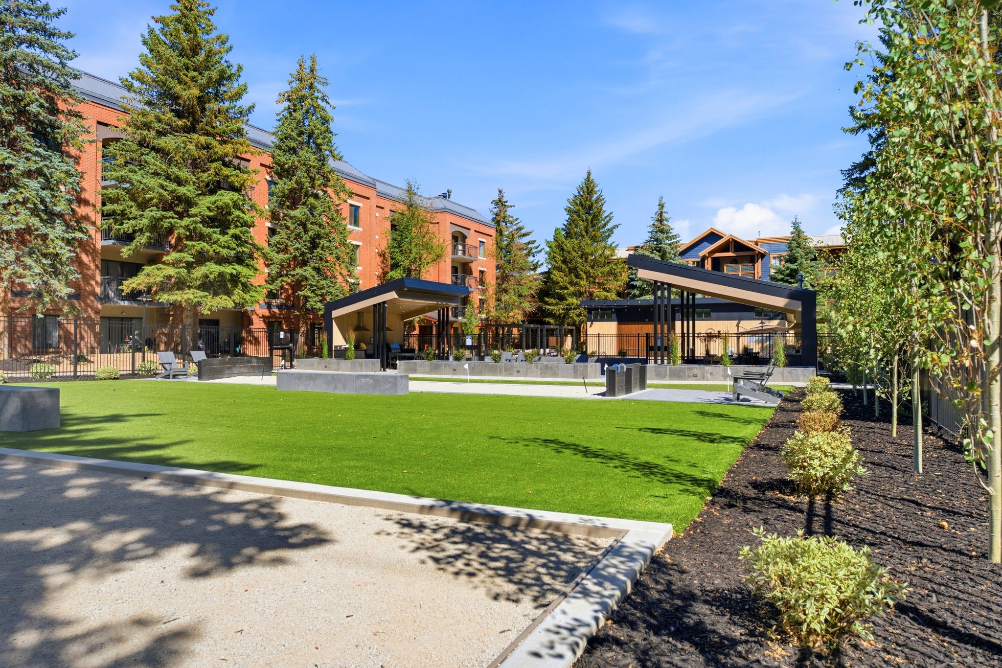 Expansive green space featuring artificial turf, bordered by mature evergreen trees and contemporary pergolas