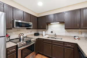 Kitchen featuring stainless steel appliances, light stone counters, dark wood finish cabinetry, and tasteful backsplash