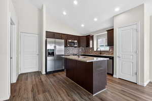 Kitchen with dark wood finish cabinetry, stainless steel appliances, vaulted ceiling, a kitchen island with sink, and light stone countertops