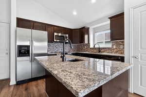 Kitchen featuring dark wood finish cabinetry, stainless steel appliances, vaulted ceiling, light stone countertops, and a center island with sink