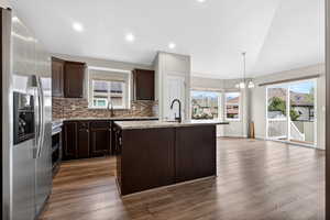 Kitchen featuring stainless steel appliances, dark wood finish cabinets, a kitchen island with sink, hanging lights, and light stone counters