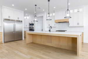 Kitchen with built in appliances, glass insert cabinets, a center island with sink, light wood-style flooring, and two tone cabinetry