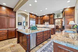 Kitchen featuring arched walkways, an island with sink, recessed lighting, built in appliances, and light stone countertops