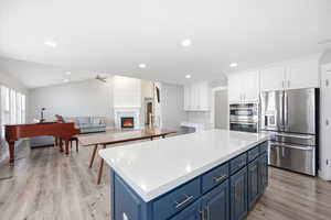 Kitchen with open floor plan, two tone cabinetry, stainless steel appliances, a warm lit fireplace, and light wood-style floors