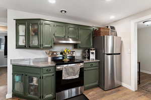 Kitchen featuring green cabinetry, glass insert cabinets, stainless steel appliances, light stone countertops, and light wood-type flooring