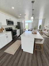 Kitchen featuring stainless steel appliances, a textured ceiling, decorative light fixtures, a kitchen island with sink, and dark wood-style flooring