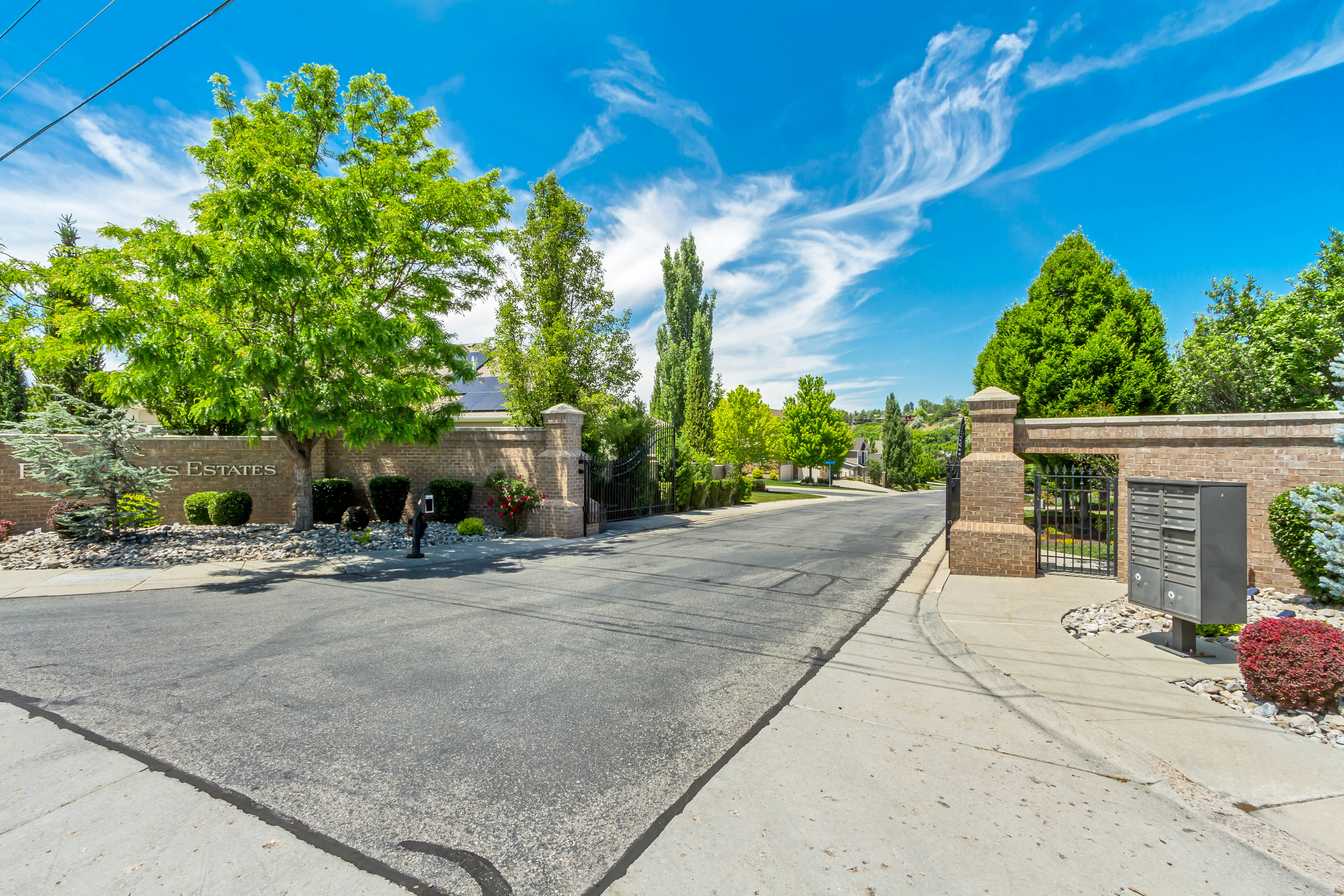 Brick entrance walls with wrought iron gates, mature landscaping, and an asphalt road