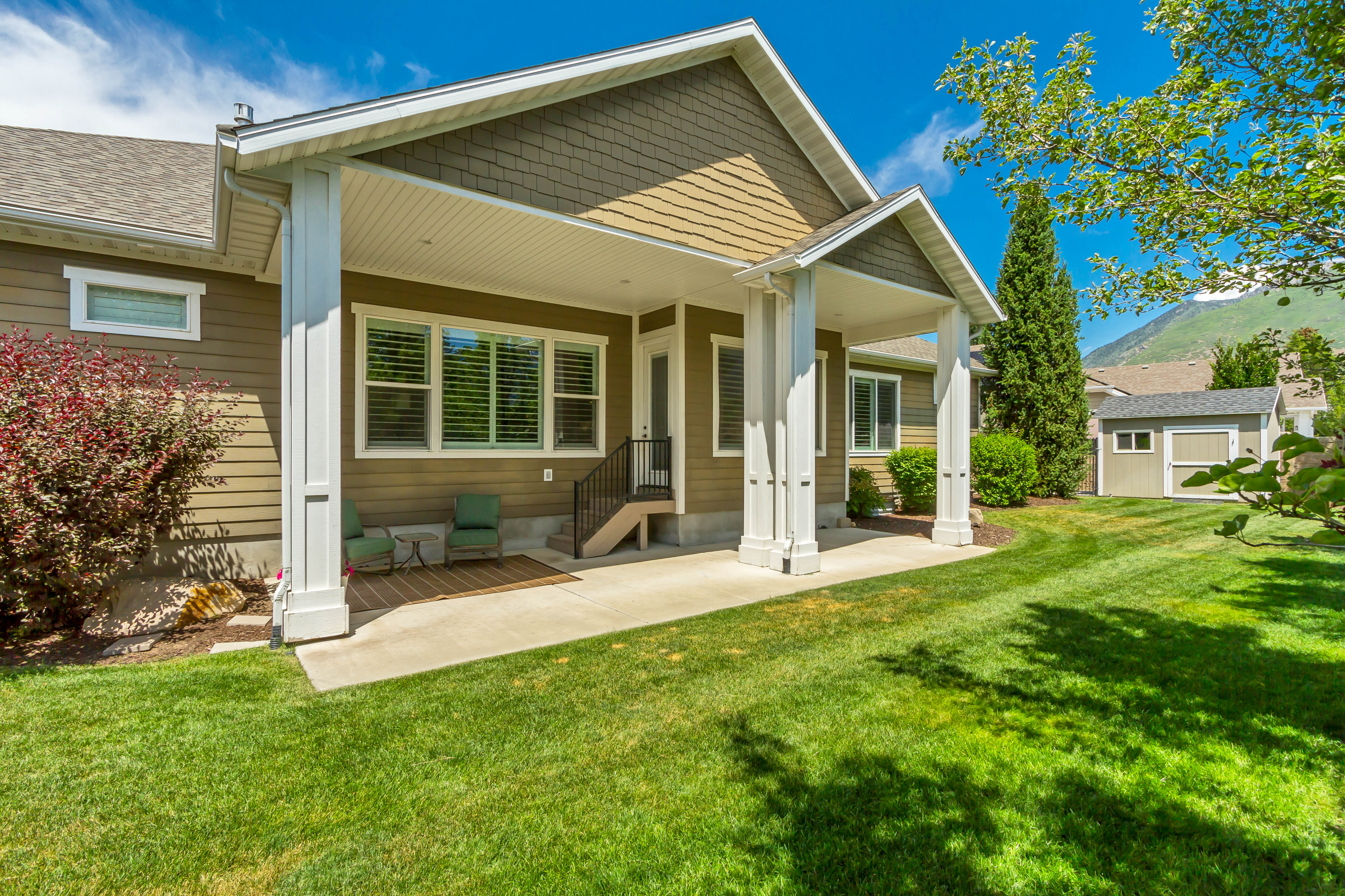 Expansive lawn with a covered patio, featuring substantial white support columns, a gray shingle roof, and a visible mountain backdrop