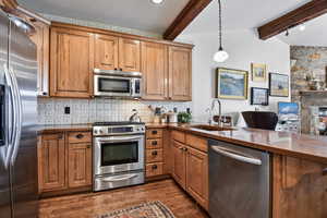 Kitchen featuring stainless steel appliances, tasteful backsplash, lofted ceiling with beams, wood finish cabinets, and dark wood-style flooring