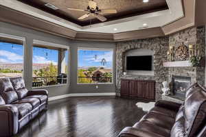 Living room featuring a ceiling fan, dark wood-type flooring, a stone fireplace, and a tray ceiling