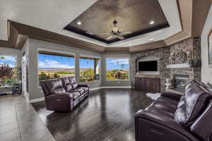 Living area featuring a ceiling fan, a stone fireplace, wood finished floors, a tray ceiling, and recessed lighting