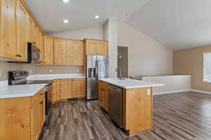 Kitchen with stainless steel appliances, light wood finish cabinets, an island with sink, dark wood finished floors, and vaulted ceiling