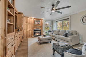 Living room featuring crown molding, light wood finished floors, a glass covered fireplace, a textured ceiling, and ceiling fan