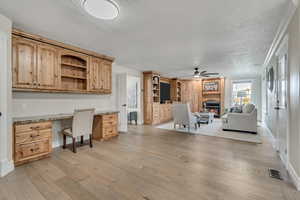 Home office with a glass covered fireplace, a textured ceiling, built in desk, light wood-type flooring, and ceiling fan