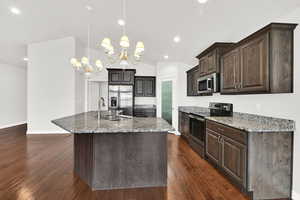 Kitchen featuring stainless steel appliances, dark wood finish cabinets, light stone counters, a kitchen island with sink, and vaulted ceiling