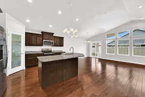 Kitchen featuring hanging lights, dark wood finish cabinets, stainless steel appliances, an island with sink, and light stone counters