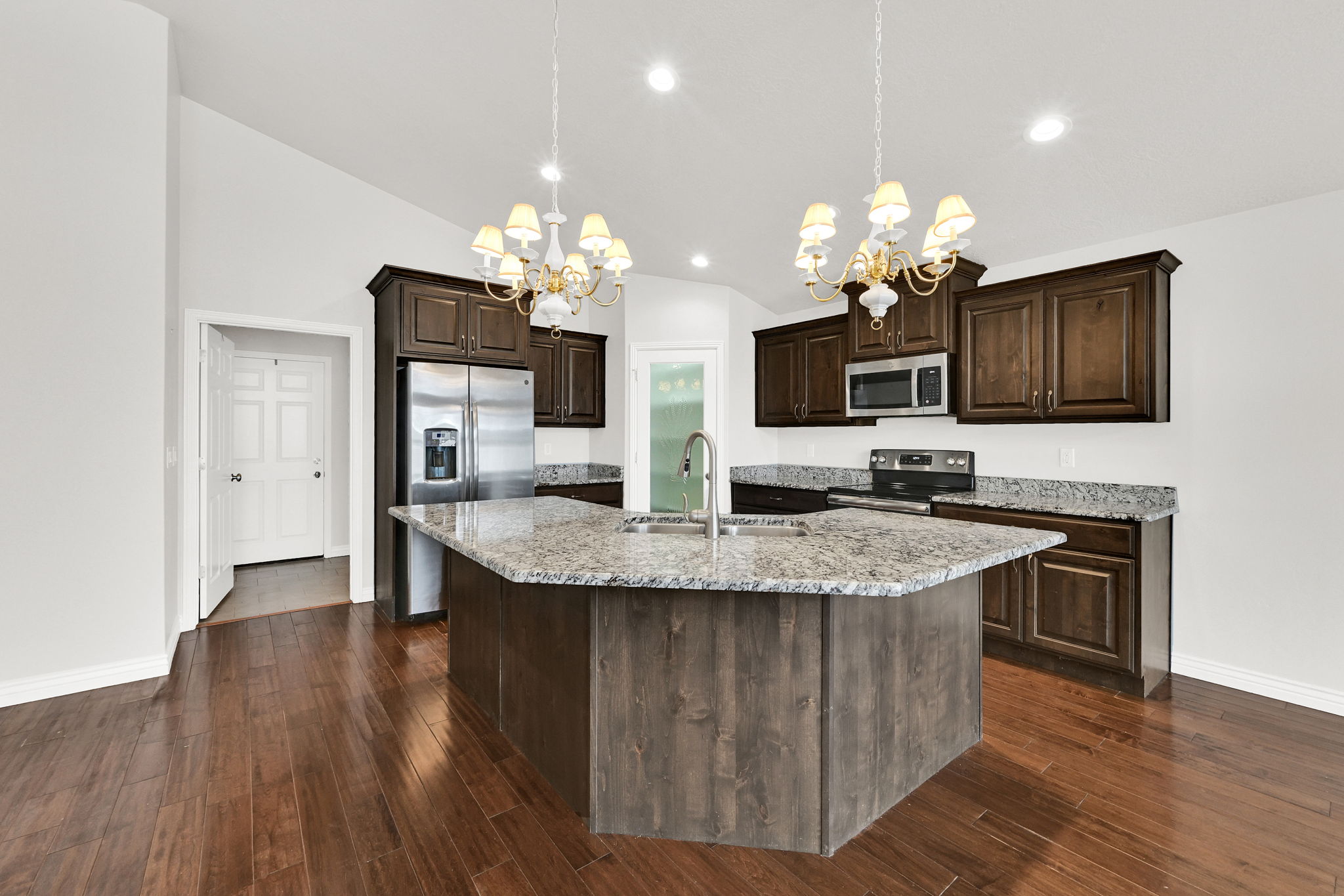 Kitchen with dark wood finish cabinets, light stone counters, a chandelier, a center island with sink, and stainless steel appliances