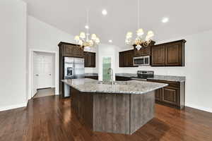 Kitchen with dark wood finish cabinets, light stone counters, a chandelier, a center island with sink, and stainless steel appliances