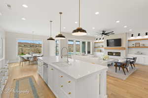 Kitchen featuring white cabinets, a large fireplace, light wood-style flooring, an island with sink, and decorative light fixtures