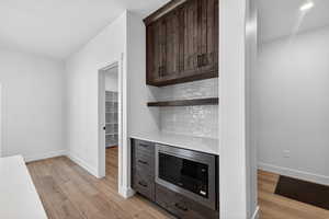 Bar area with stainless steel microwave, dark wood finish cabinets, light wood-style floors, tasteful backsplash, and light stone counters