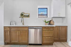 Bar featuring stainless steel dishwasher, light stone counters, light wood-style floors, and two tone color scheme