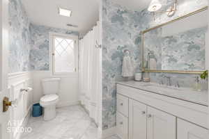 Full bathroom featuring wallpapered walls, vanity, a shower with curtain, a wainscoted wall, and light marble finish flooring