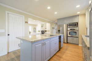 Two tone kitchen with stainless steel appliances, a center island, light stone countertops, light wood finished floors, and ornamental molding