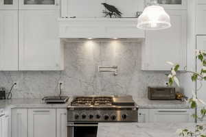 Kitchen with gas range, white cabinetry, light stone countertops, glass insert cabinets, and decorative backsplash