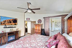 Bedroom featuring a barn door, ceiling fan, light carpet, ensuite bath, and a glass covered fireplace