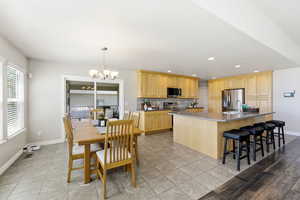 Kitchen featuring light wood finish cabinetry, a center island with sink, stainless steel appliances, light tile patterned flooring, and hanging lights