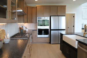Kitchen featuring stainless steel appliances, dark stone countertops, light wood-style flooring, glass fronted cabinets, and tasteful backsplash