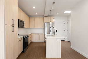 Kitchen with a center island with sink, light wood finish cabinetry, stainless steel appliances, and dark wood-type flooring