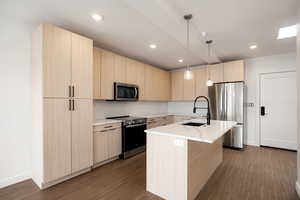 Kitchen with light wood finish cabinetry, stainless steel appliances, an island with sink, and dark wood-type flooring