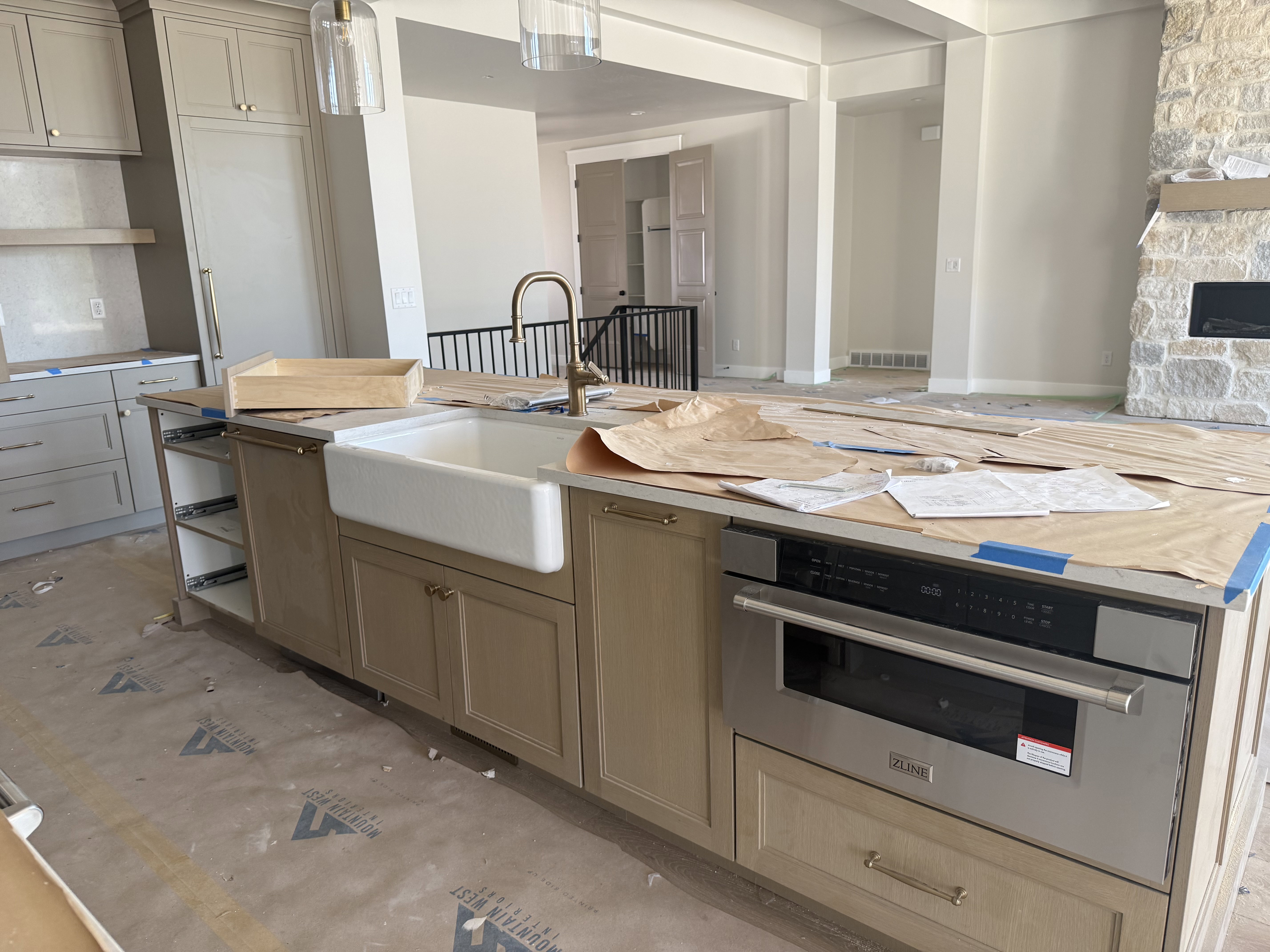 Kitchen island with a farmhouse sink, stainless steel built-in microwave, light-toned cabinetry, gold-finish hardware, and a stone-clad fireplace in the background