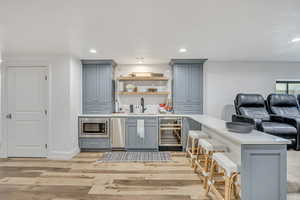Bar area with gray cabinets, beverage cooler, light stone counters, stainless steel microwave, and light wood-type flooring
