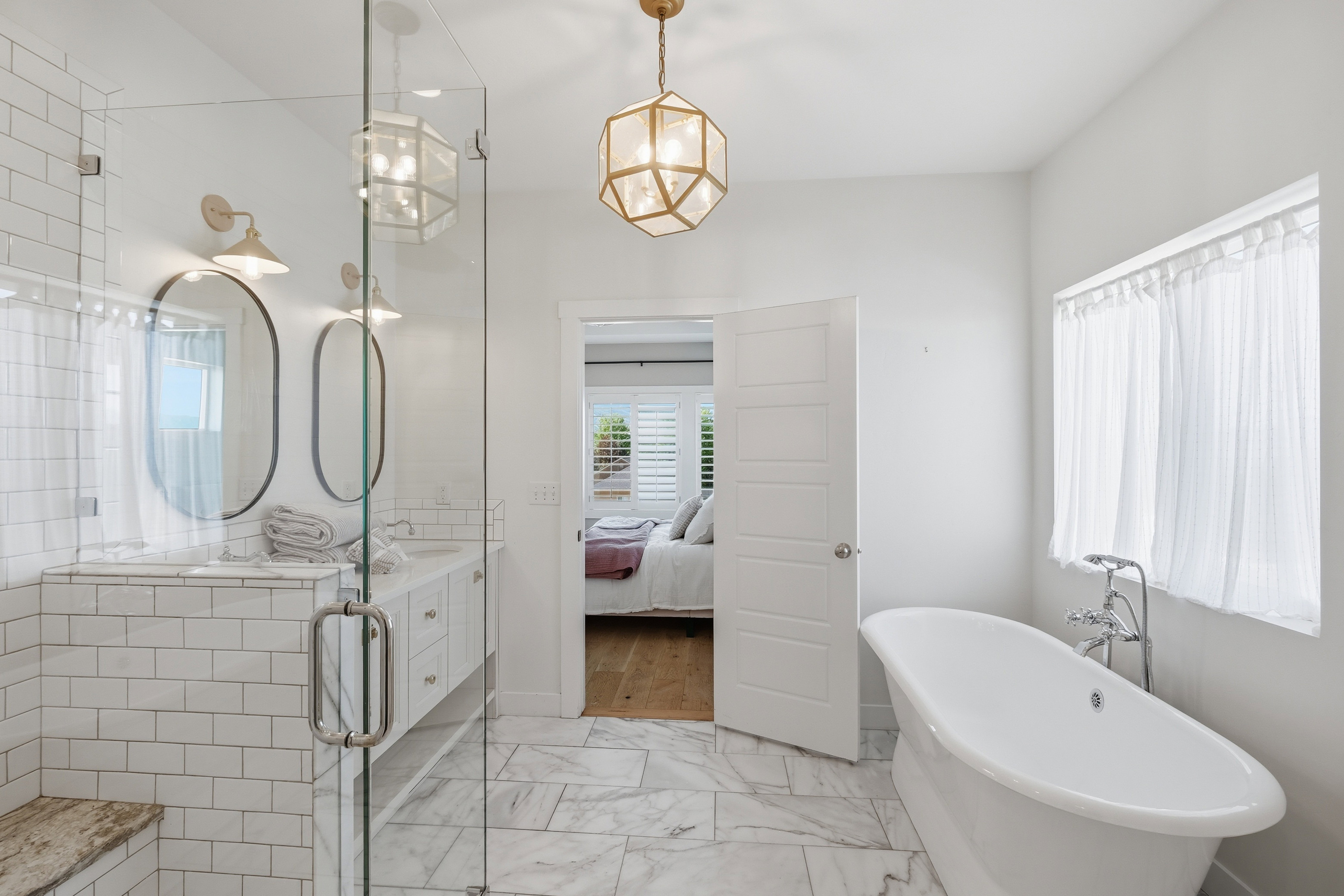 Spacious bathroom featuring a freestanding soaking tub, large walk-in shower with a built-in bench, double vanity with dual oval mirrors, marble-look tile flooring, and a unique geometric pendant light fixture