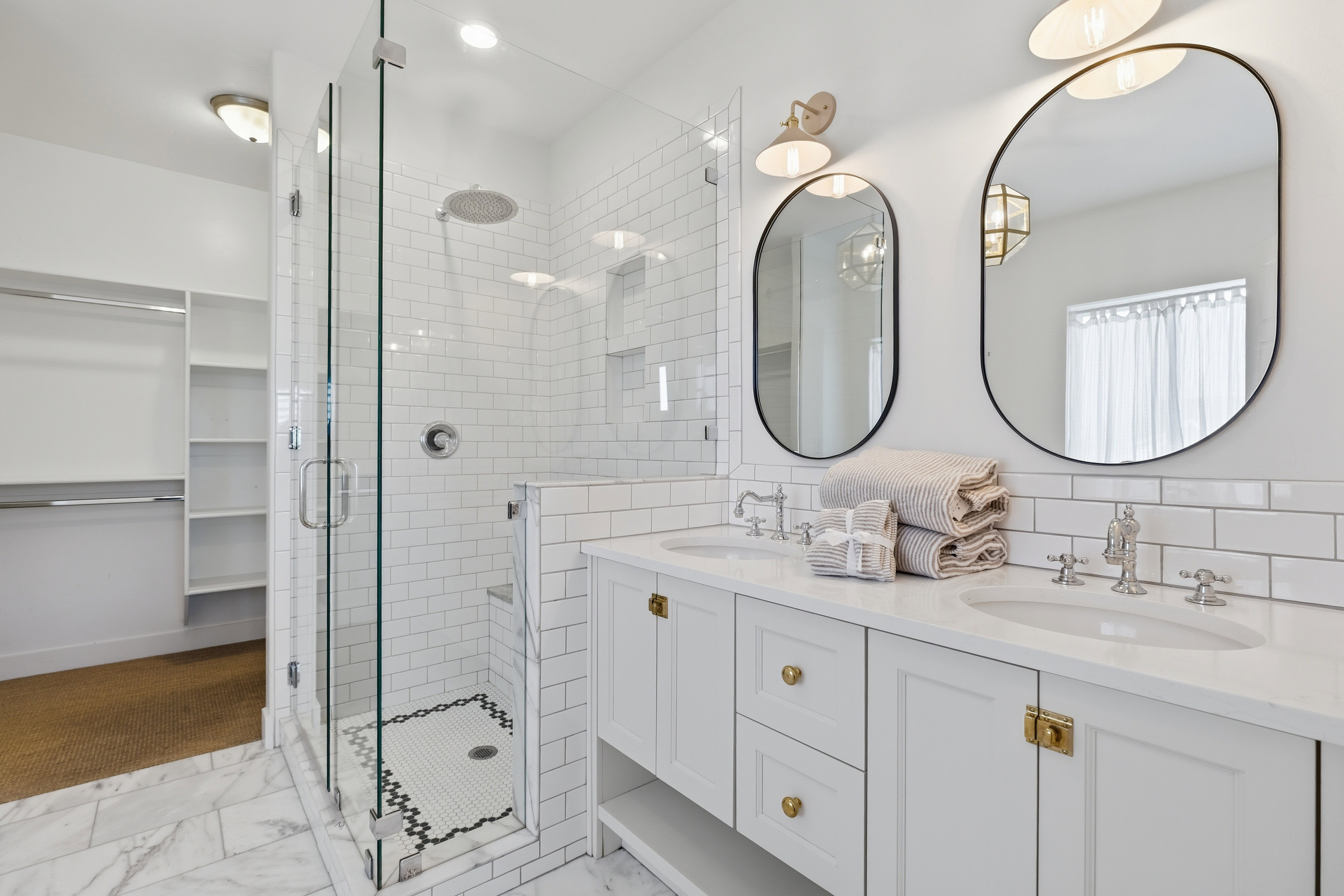 Bathroom featuring a dual vanity with white cabinetry and brass hardware, two oval framed mirrors, and chrome fixtures