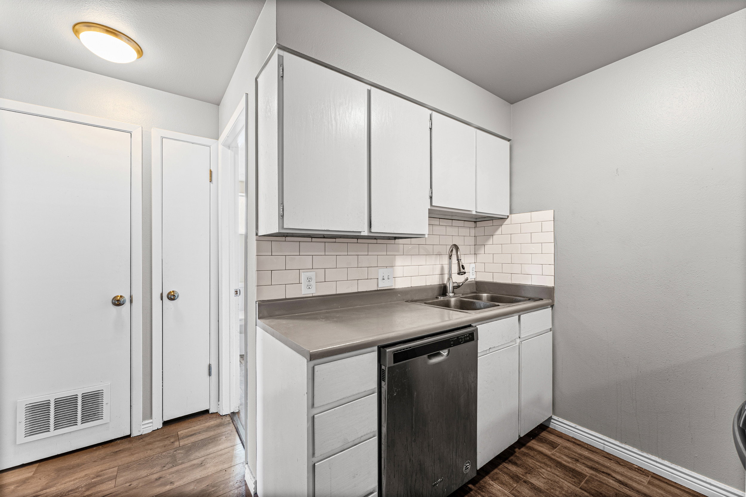 Kitchen with stainless steel dishwasher, decorative backsplash, dark wood-style floors, white cabinets, and light countertops